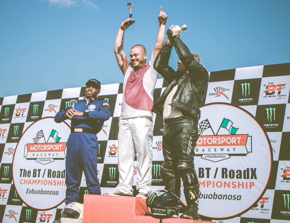 Three race car drivers standing on a podium with hands in the sky and a small trophies.