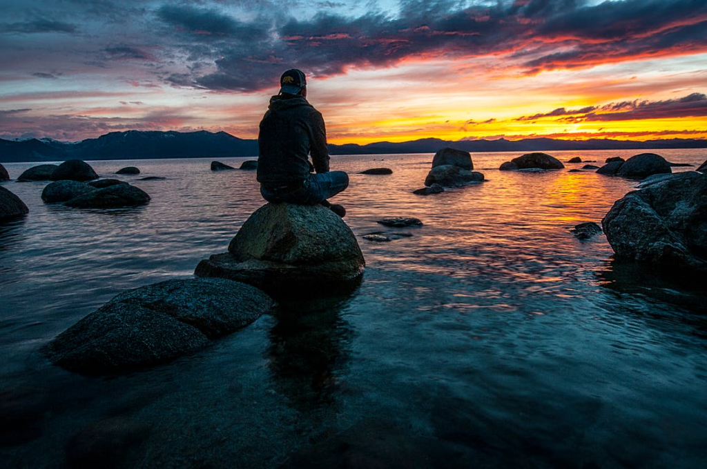 Young man with backwards hat sitting on a rock by the sea shore, watching a beautiful sunset in the horizon.