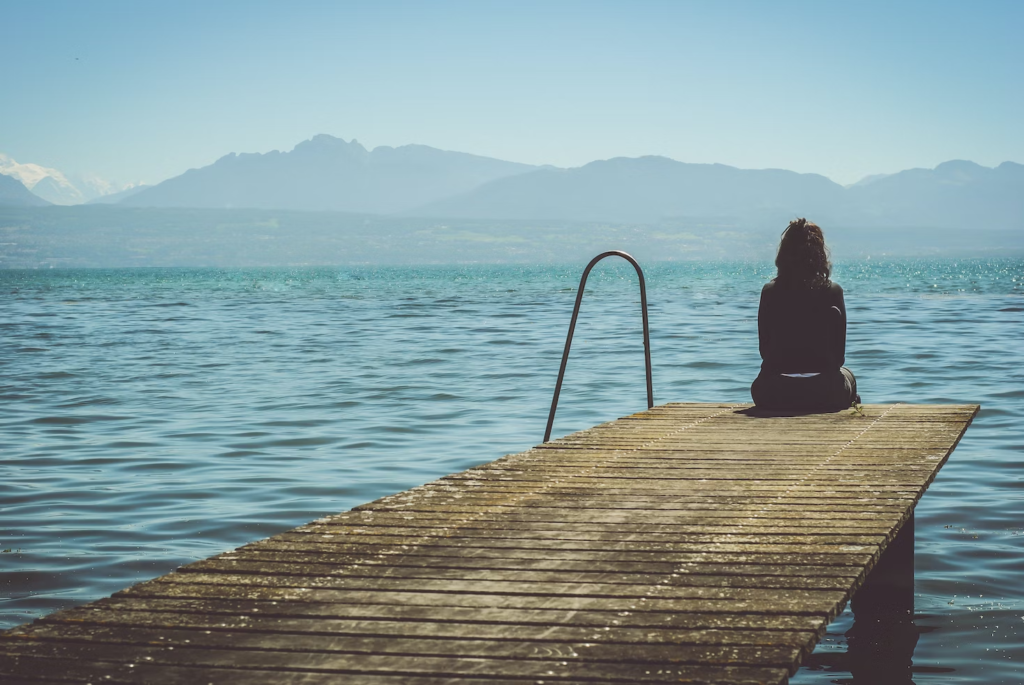 Woman sitting alone on a dock in solitude, overlooking the sea and mountains.