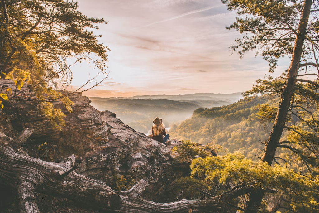 Woman with hat sitting in nature on a cliff overlooking a lush, green landscape.